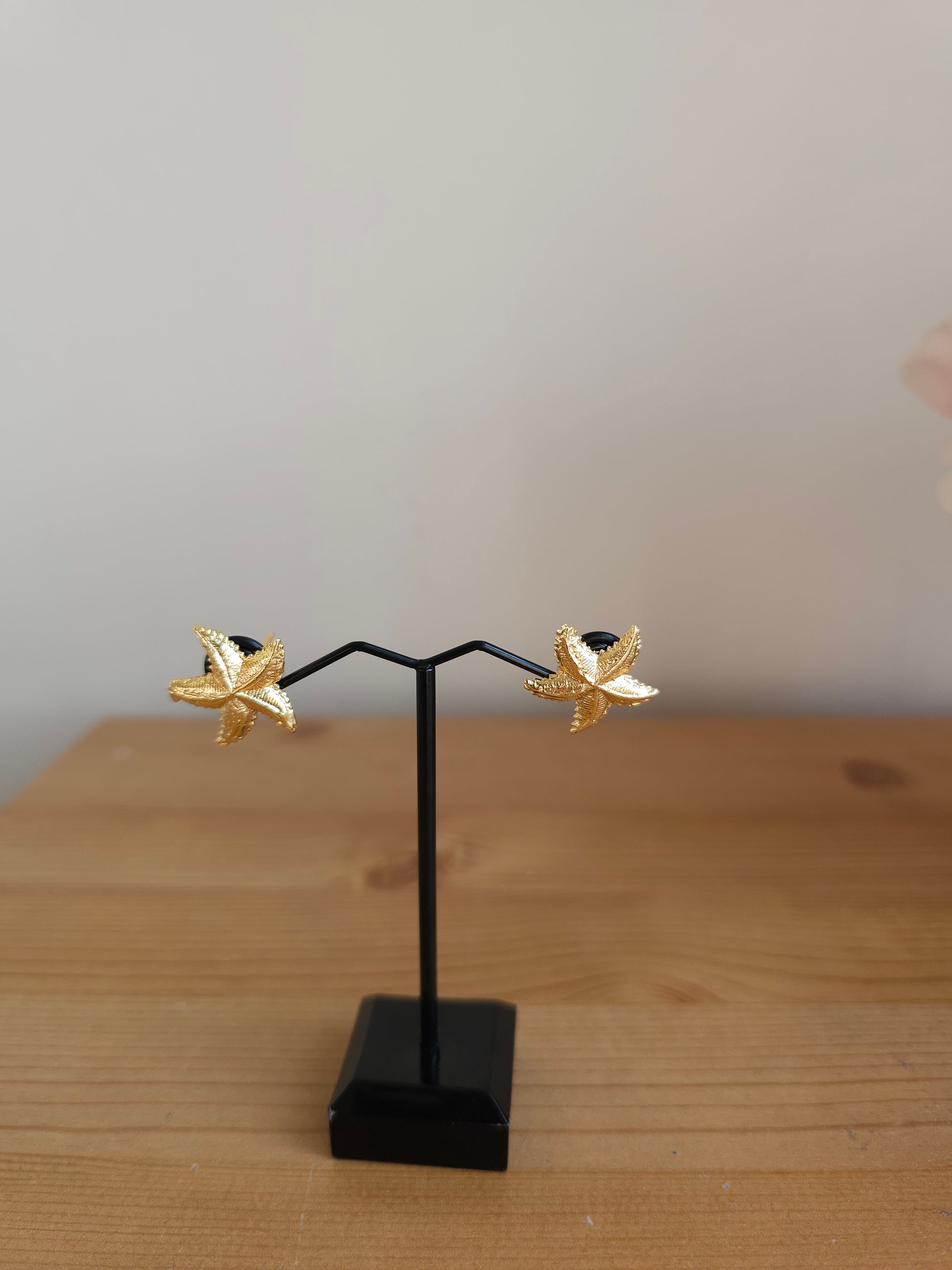 Gold flower-shaped earrings on a black stand against a wooden background